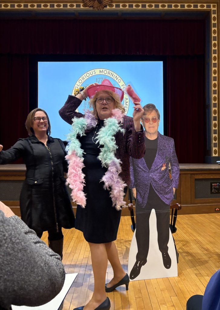 Retiring Select Board member Suzie Barry poses in colorful garb next to Cary Library Director Koren Stembridge and a cardboard cutout of Elton John